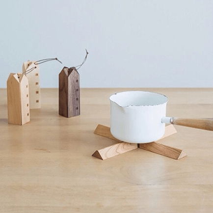 写真：白い片手鍋が置かれた木製のクロス型鍋敷き。畳むと家の形のオブジェになる。